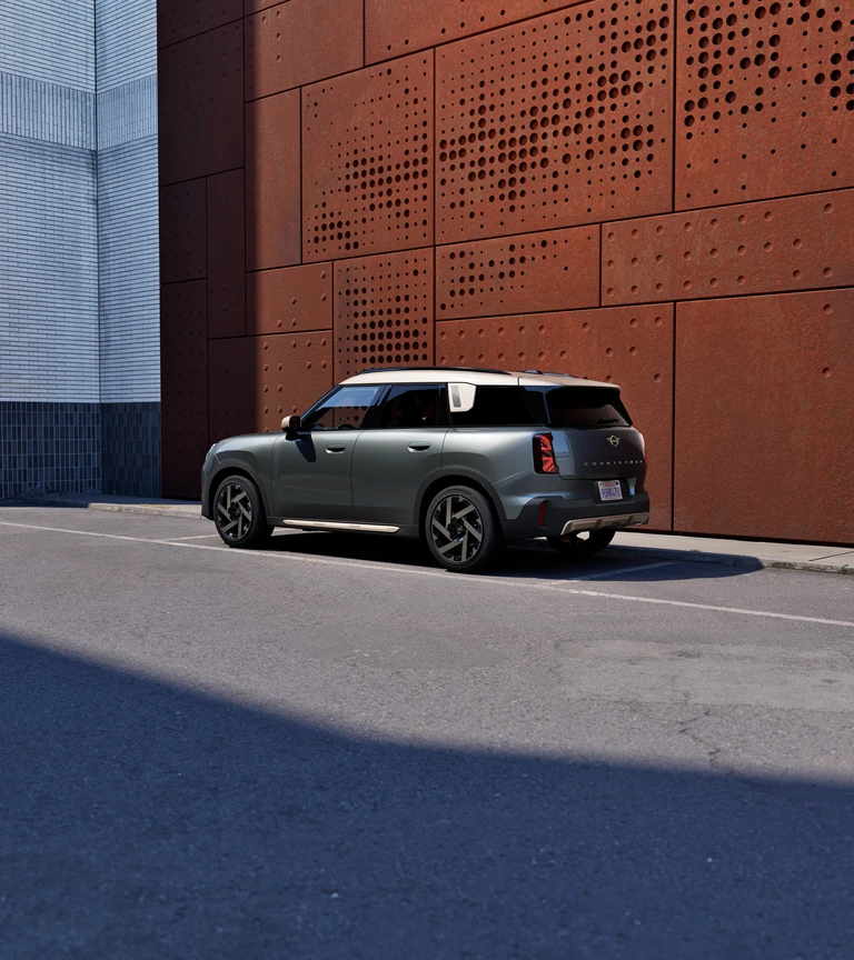 Rear three‑quarter view of a MINI Countryman in Sage Green Metallic parked beside a perforated rust‑coloured wall.