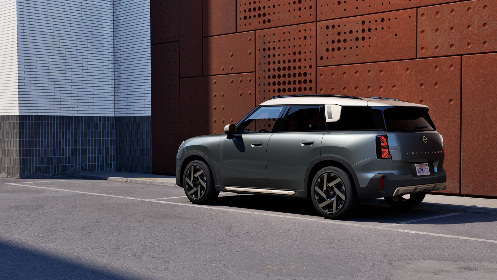 Rear three‑quarter view of a MINI Countryman in Sage Green Metallic parked beside a perforated rust‑coloured wall.