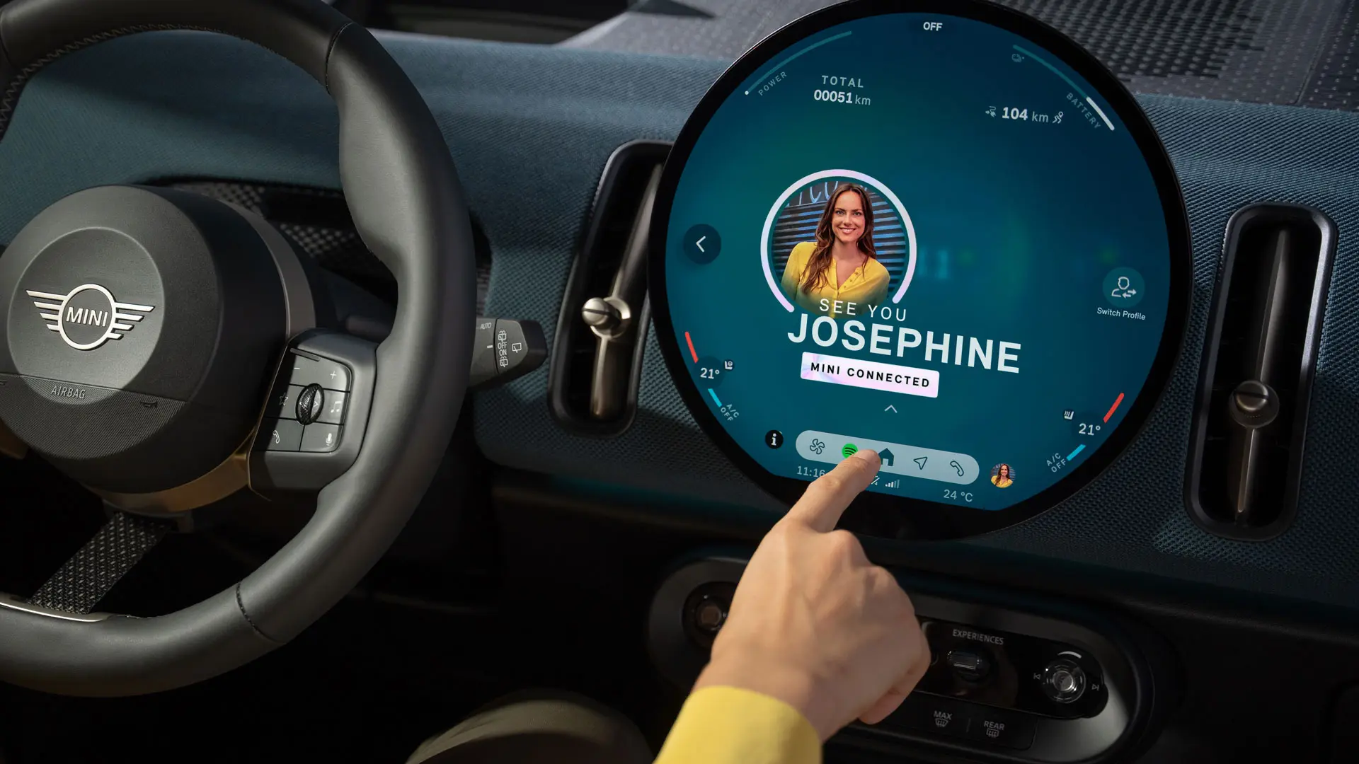 Driver interacting with the circular MINI infotainment display showing a MINI Connected profile screen.