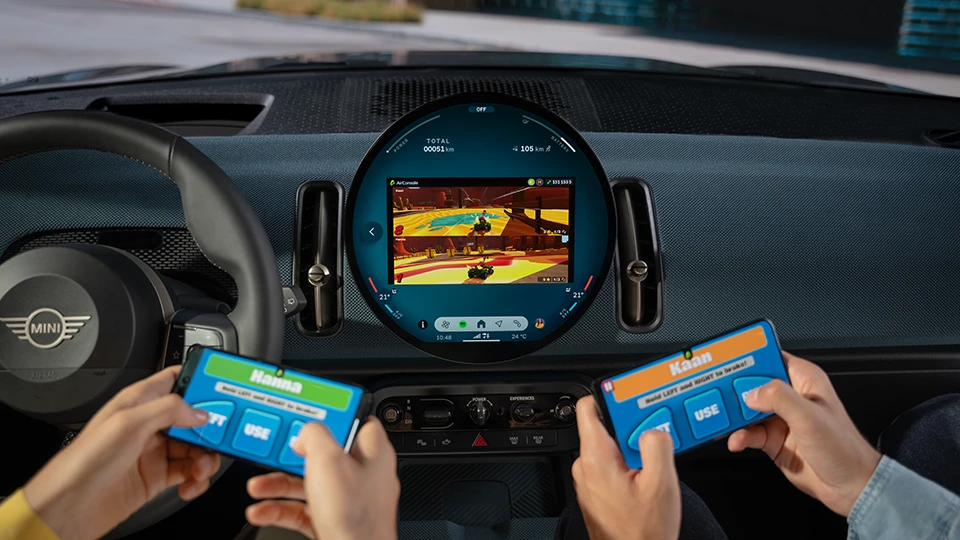 Two people using handheld controllers to play a game displayed on the MINI circular infotainment screen inside the vehicle.
